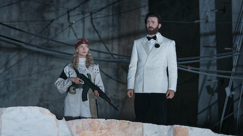 sacrifice-netflix-chris-evans-anya-taylor-joy-october-2026-cover A woman in an adventure outfit stands beside a bearded man in a white tuxedo in a Netflix still from Sacrifice